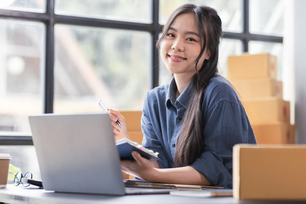 An Asian girl smiling while holding a notebook in front of her laptop